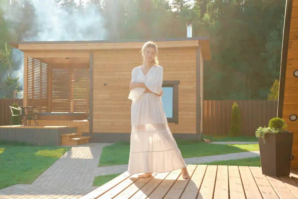 Verträumte Frau in langem weißem Kleid steht auf Terrasse mit Whirlpool und Garten-Sauna aus Holz im Hintergrund - Entspannung pur im eigenen Garten: Tipps für den Kauf einer Gartensauna aus Holz 